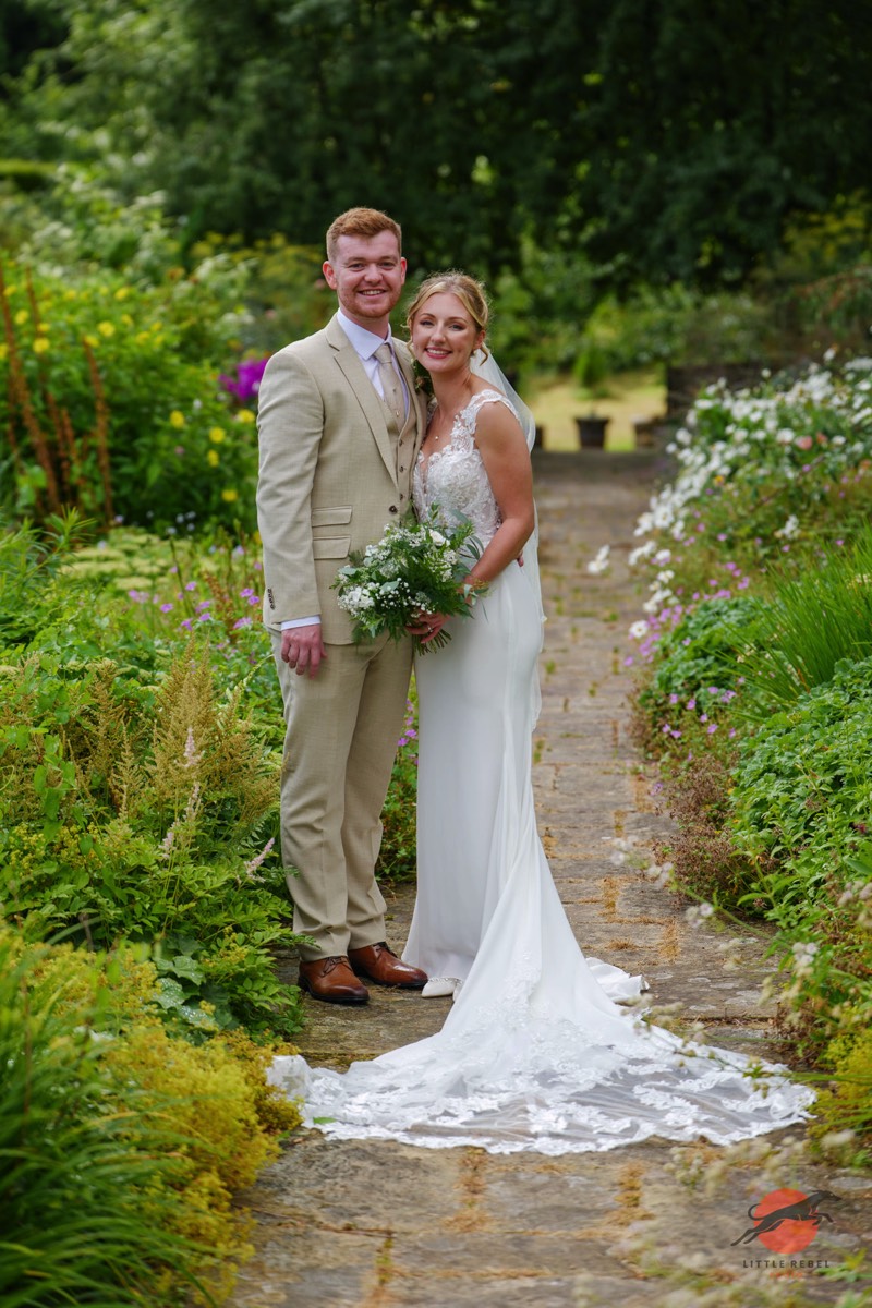 Newlyweds standing together on a flower-lined garden path at Askham Hall