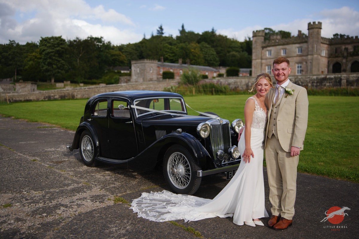Newlyweds beside a vintage MG VA wedding car outside Lowther Castle in Cumbria